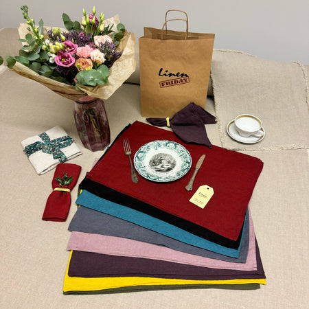 Linen placemats of different colors, beg with LinenFriday logo, vase with flowers, plate, fork and knife, cup and little plate.