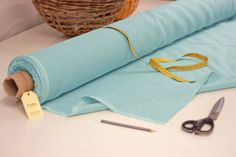 mint blue linen fabric roll, scissors and basket on white table