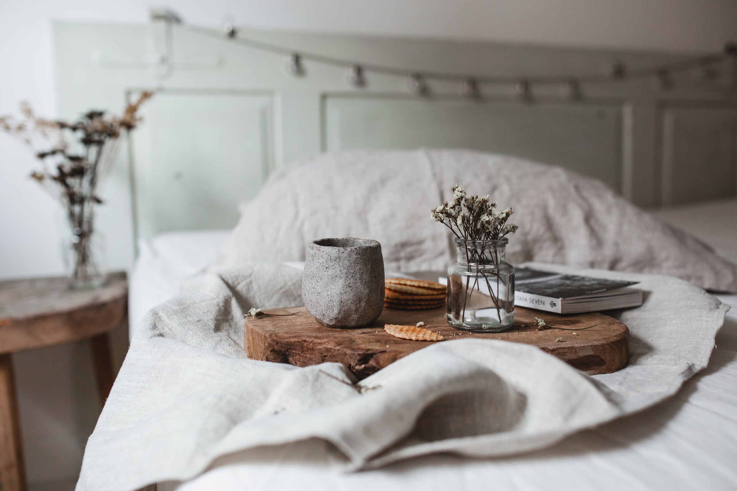 A room with a wooden table and a vase of flowers, a bed with light linens, on which there is a wooden tray with a book, jars and crumbled cookies